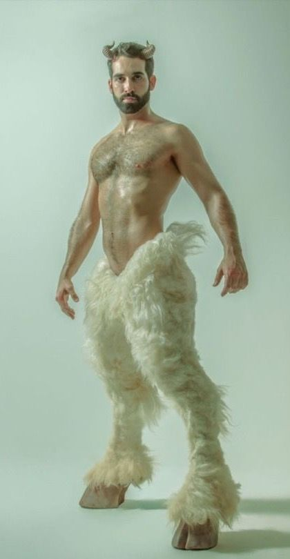 a full body shot of a male model posing as a satyr.  he's shirtless, with a hairy chest, and wears a costume consisting of goat legs made of light creamcolored fur and small horns on his forehead. the background is a pale, light green. the image has an artistic and slightly surreal quality.