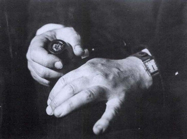 the black and white closeup photograph shows the hands of an older man.  his left hand holds a pipe, likely a tobacco pipe, showing the bowl and the stem. his right hand rests on his lap, revealing a classicstyle wristwatch. the image has a grainy texture characteristic of older film photography. the lighting creates strong contrasts and shadows, emphasizing the wrinkles and texture of the man's skin. the overall mood is somber and reflective. the focus is sharply on the hands and the pipe, while the background remains blurred and indistinct.