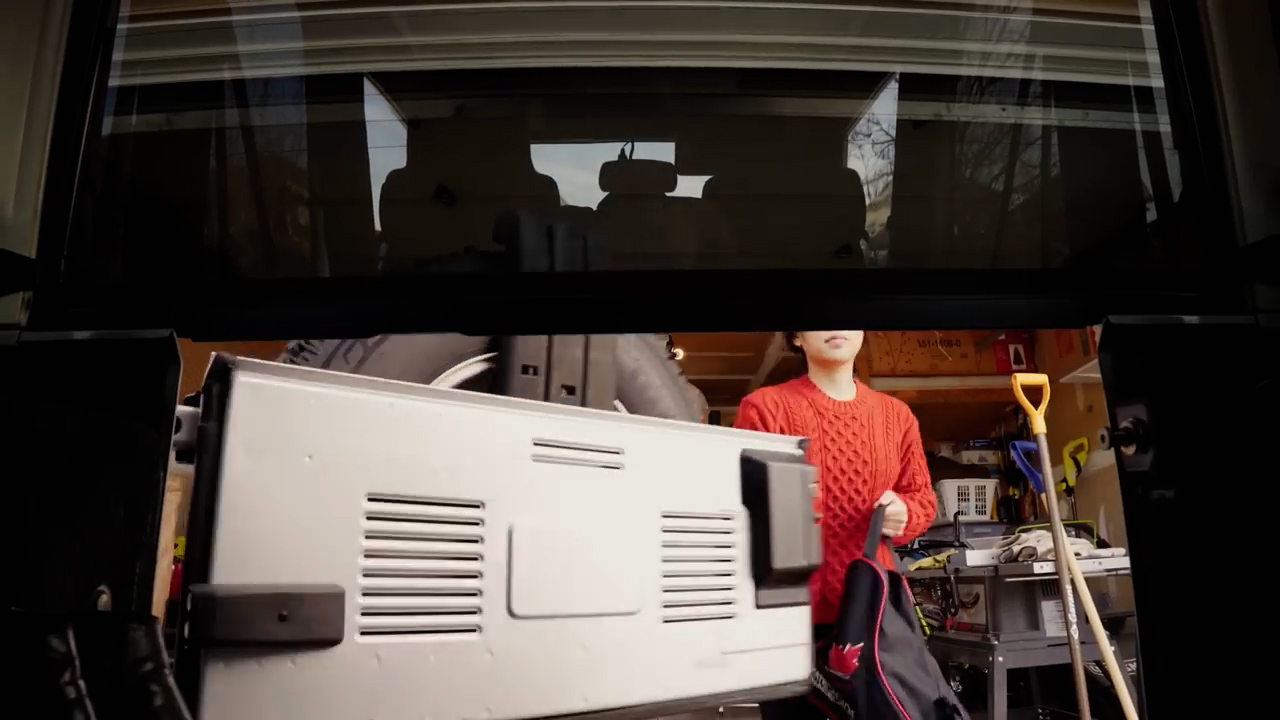 a man in a red sweater is loading luggage into the back of a car. the image is taken from inside the trunk, looking out. the man is standing in a garage, and the car's back window is open.  the luggage is a silver hardshell suitcase. there is a red backpack on the ground to the right of the suitcase.  the garage is cluttered with tools and other items.