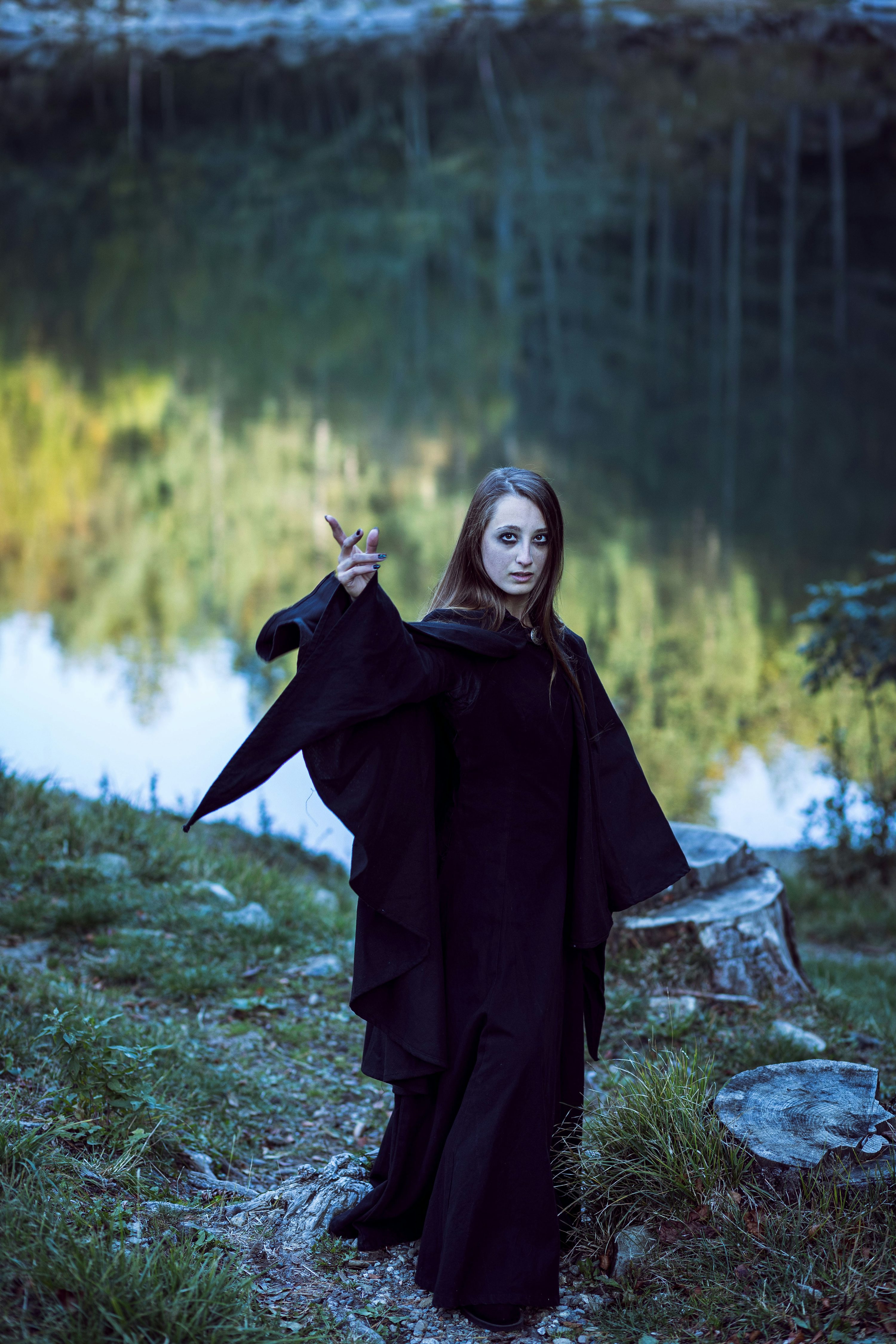 a woman dressed in a long, dark hooded cloak stands by a lake in a forest. she appears to be casting a spell, with one hand raised in a gesture. the scene is moody and atmospheric, with a mystical and supernatural feel.  the woman's pale skin and dark clothing contrast with the green and blue tones of the natural setting. the image suggests a fantasy or gothic aesthetic.