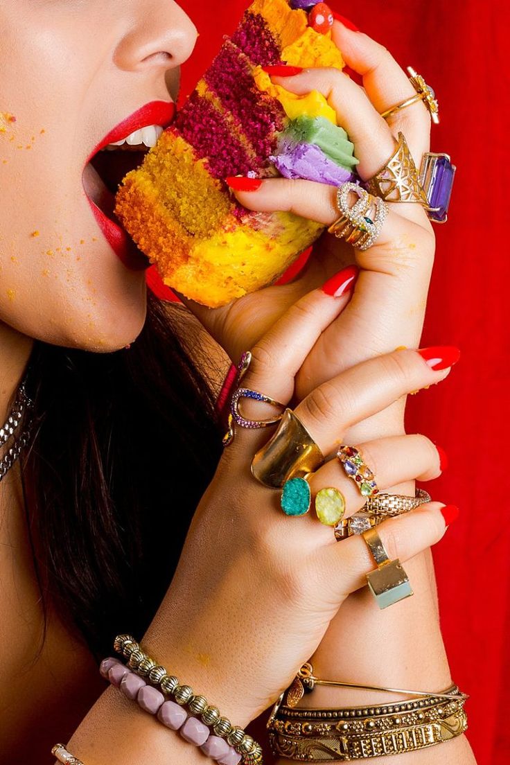 closeup view of a woman with long dark hair and red nails biting into a vibrant, layered rainbow cake. she is adorned with numerous gold rings featuring various gemstones and multiple bracelets and bangles on both wrists. the background is a solid, deep red, providing a striking contrast to the colorful cake and the woman's jewelry. the image is rich in color and detail, focusing on the textures and vibrancy of the food and accessories.
