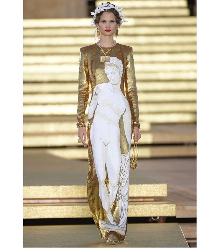 a model wearing a gold dress with a white statue print is walking on a runway. the dress has long sleeves and is floorlength. the model is wearing a necklace, earrings, and a headdress. the runway is gold and has a stage in the background.
