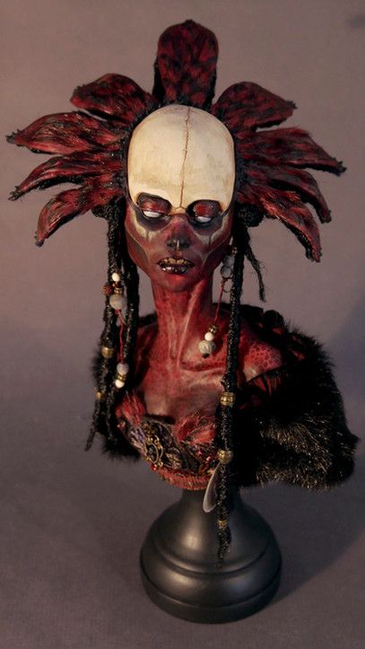this is a closeup image of a doll or figurine sculpted to resemble a female creature with red feather headdress, long black hair, and red skin. the doll's face is painted with exaggerated white features and the figure is wearing a dark costume with a red chest piece. it is mounted on a wooden stand and is posed against a plain gray background.