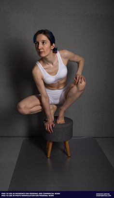a woman in white athletic wear poses balanced on a small stool in front of a gray backdrop in a studio setting. she is holding the stool with her right hand for balance. the lighting is soft and diffused.