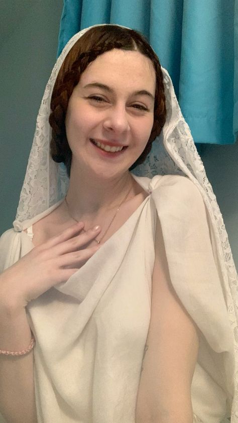 a young woman smiles at the camera, dressed in a white historicalinspired costume. she wears a white lace veil over her head and a draped white dress. her hair is styled in a braided updo. the background is a teal curtain and a light blue wall, suggesting an indoor setting. the lighting is artificial and the perspective is at eyelevel.