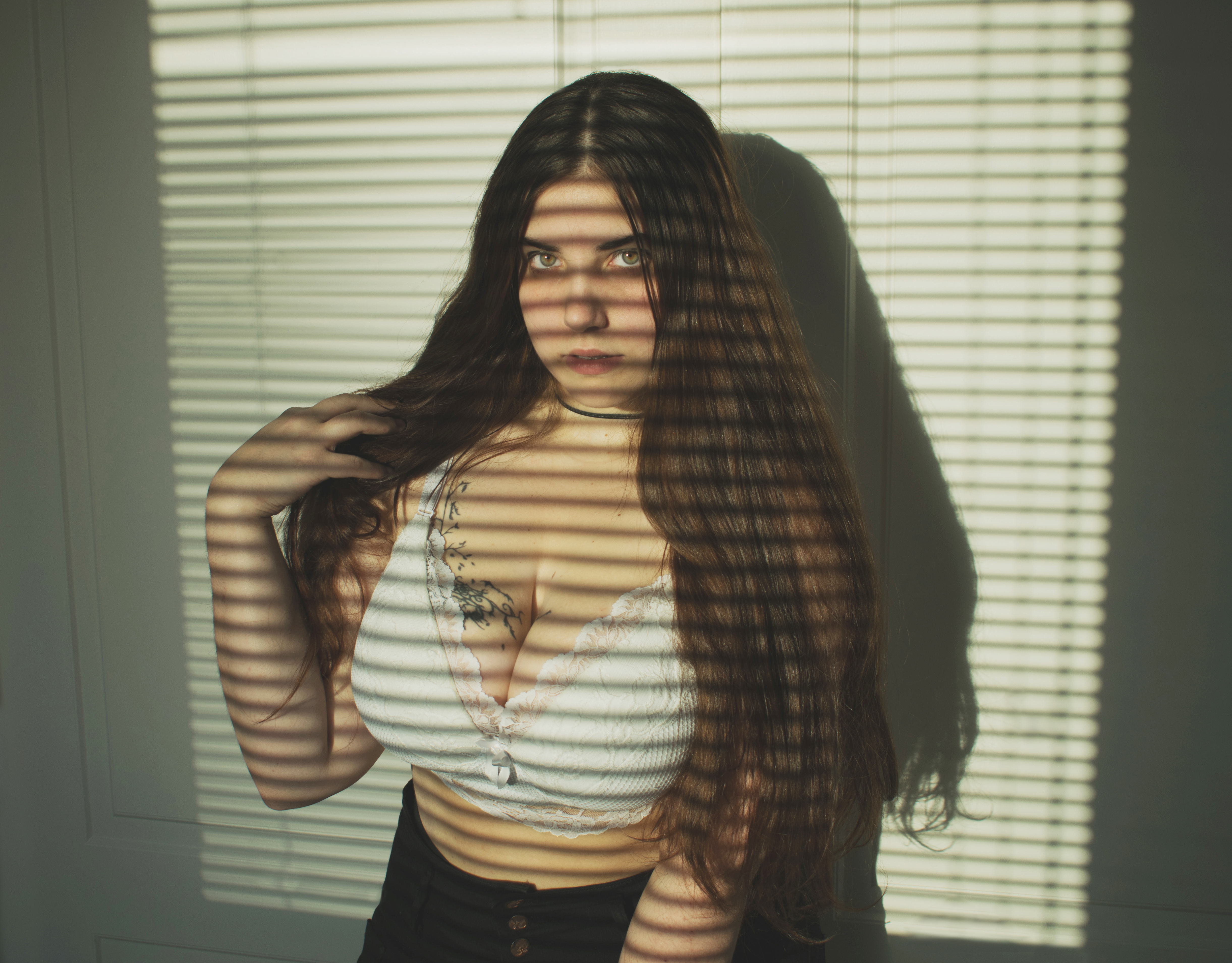 a woman with long brown hair stands in front of window blinds, her body partially obscured by the shadows cast by the blinds. she is wearing a white lace top and black shorts. she has a tattoo on her chest and a black choker around her neck. she is looking at the camera with a serious expression. the light from the window blinds creates a striped pattern on her body and the wall behind her.