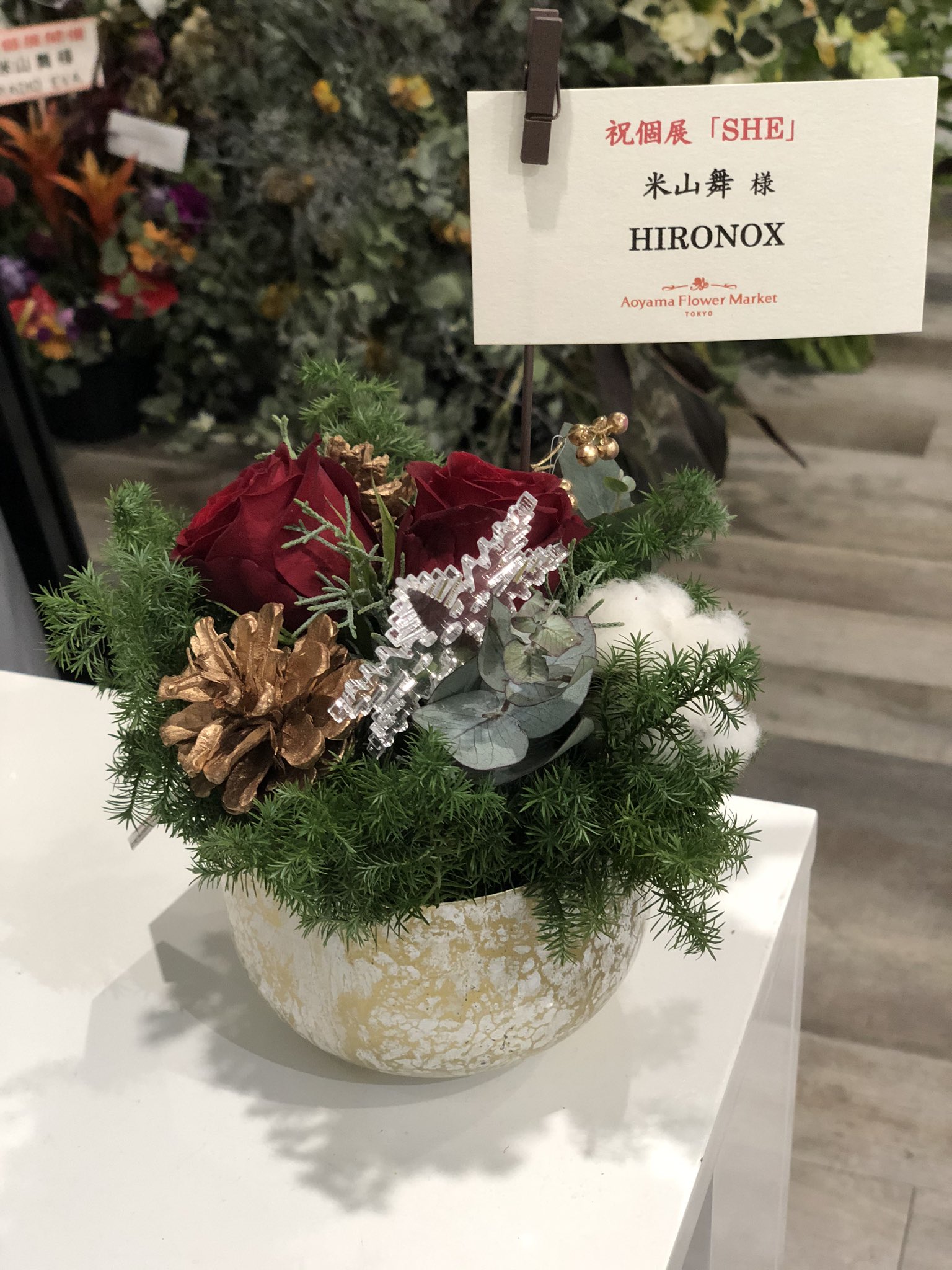 a small christmas flower arrangement with red roses, pine cones, and greenery in a gold pot sits on a white counter. there is a small sign in front of the arrangement with japanese writing on it.