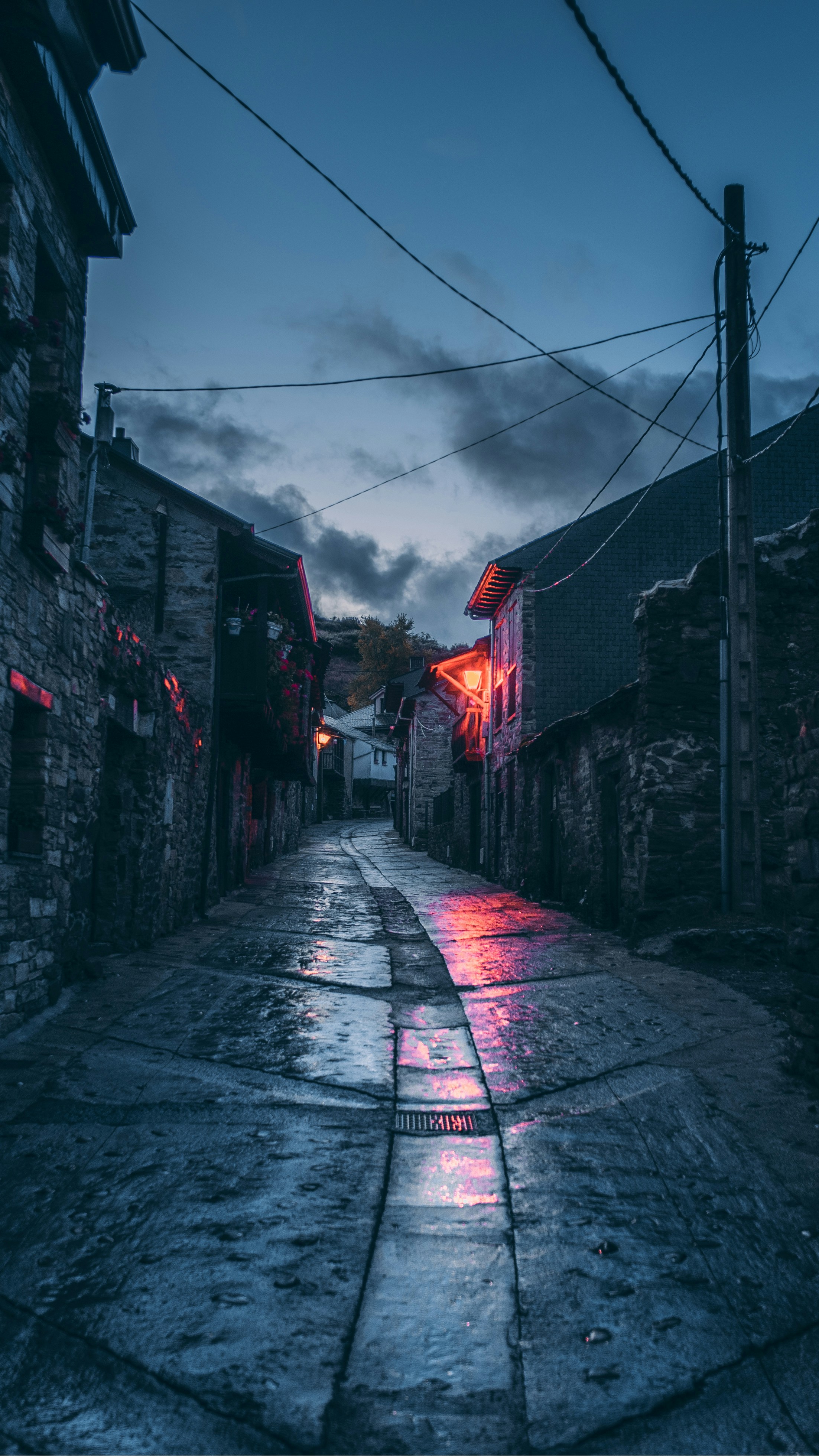 a dimly lit, narrow cobblestone street at dusk. the street is lined with old stone buildings and is wet, possibly from rain. there are no people in sight, creating an eerie and deserted atmosphere. the image evokes a sense of quiet and mystery.