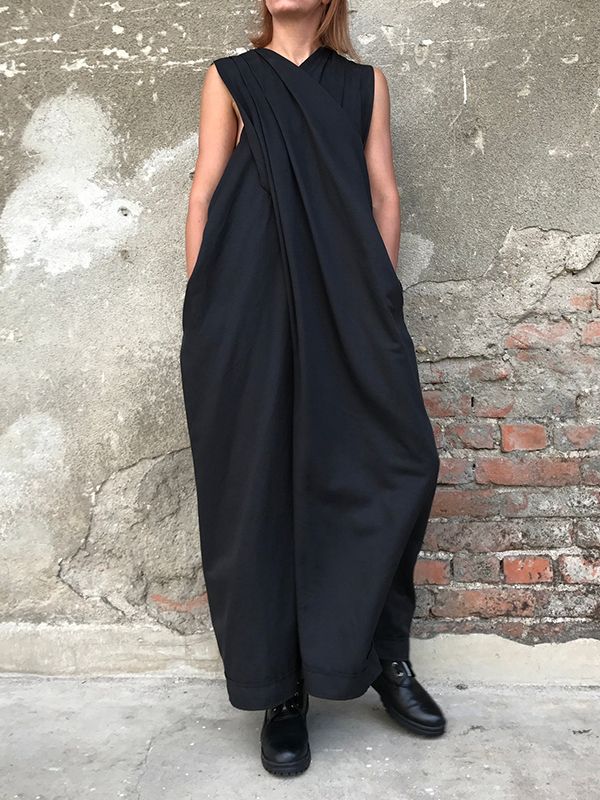 a woman in a long black sleeveless dress, with the top draped in a unique way, stands in front of a weathered brick wall, posing for a photo. she is wearing black boots.