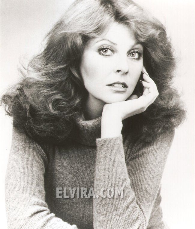 a black and white portrait of a woman with long, flowing hair. she is wearing a turtleneck sweater and has a serious expression on her face. she is looking directly at the camera.  she is posed with her hand on her chin.  her hair is styled with bangs.  she is wearing makeup.  she is in a studio setting.   the photograph appears to be from the 1970s.