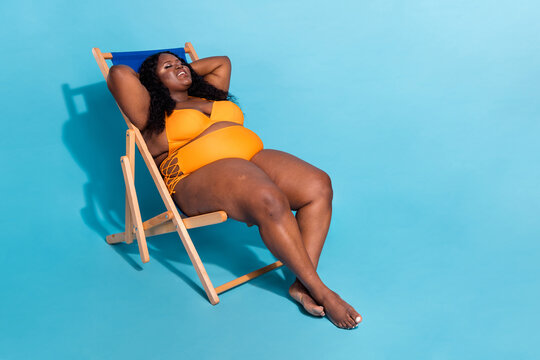 a black woman in a yellow swimsuit is relaxing in a lounge chair on a blue background. she is smiling and appears to be happy and carefree.  the chair is wooden with a blue cushion.