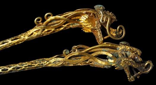 the image shows two antique gold hairpins with dragon head decorations. the hairpins are intricately designed with swirling patterns and each features a dragon head at the top, adorned with a turquoise stone. the dark background highlights the gold and details of the artifacts.