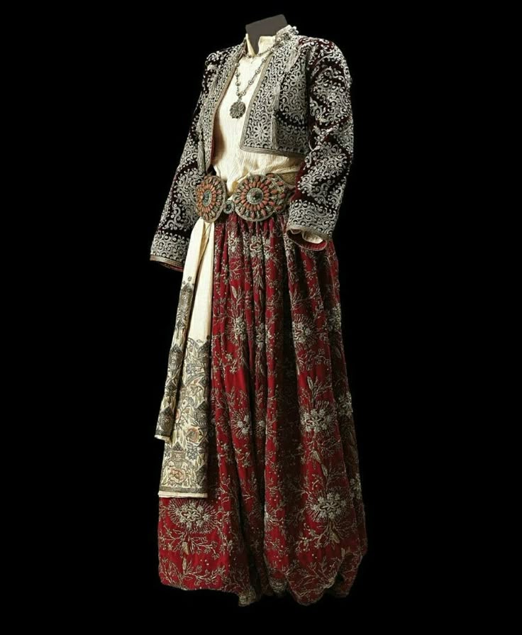 a full view of a traditional outfit consisting of a long red and white floral patterned skirt, a white blouse, a short embroidered jacket, and a decorative belt with large circular elements. the outfit also includes a draped piece of fabric and a necklace.