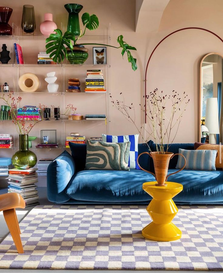 a living room with a blue sofa and a yellow coffee table, with a checkered rug in the middle of the room. the sofa is decorated with throw pillows and has a wooden armchair on the left. the room has plants and vases, and a bookshelf on the wall with several books on it. the walls are pink, and the room has a minimalist and modern decor.