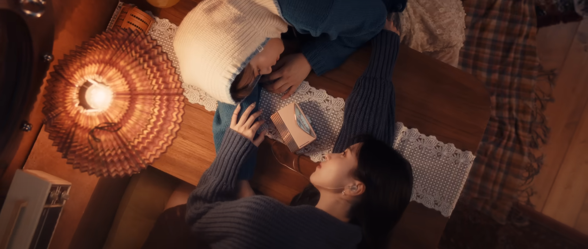 two women lie facing each other on a wooden table, their arms intertwined. the woman on the left wears a white beanie and a blue sweater, her head resting on her arm. the woman on the right wears a dark blue sweater and has her eyes closed. a small, intricately decorated box rests between them. a vintage lamp with a patterned shade hangs above, casting a warm, soft light on the scene. the environment is cozy and intimate, suggesting a loving relationship.