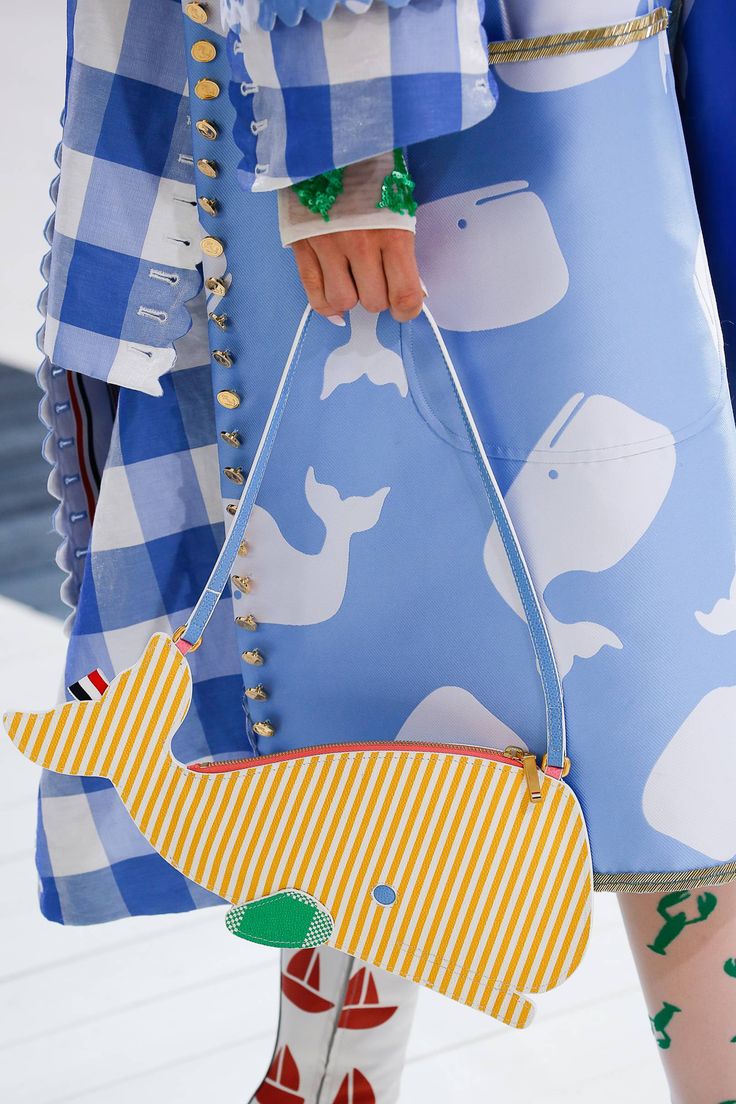 closeup shot of a model wearing a blue and white gingham dress and holding a yellow and white striped whaleshaped handbag. the handbag has a light blue strap and a small green detail on the whale's spout. the dress is decorated with white whale prints and features gold buttonlike details along the sides. the model's tights have a nautical theme with small sailboat and crab prints.  the overall style is playful and summery.