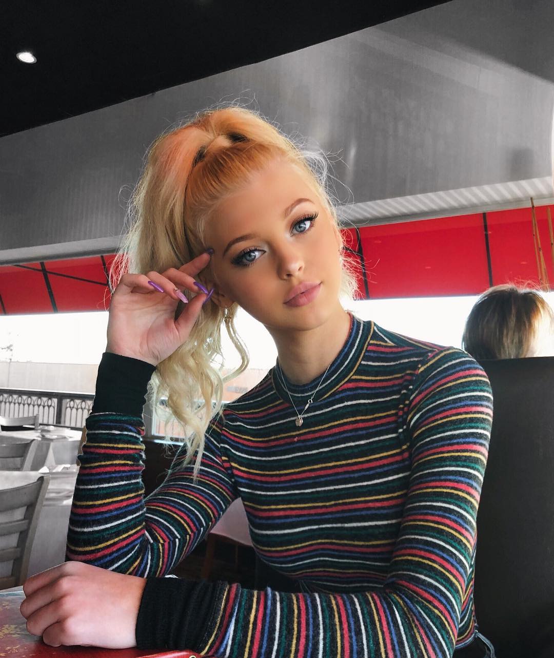 a young woman with blonde hair in a high ponytail is sitting at a table. she is wearing a longsleeved, striped shirt with black cuffs. she has on makeup and is wearing a necklace. her nails are painted a light color. she is resting her hand on her face, looking at the camera with blue eyes.