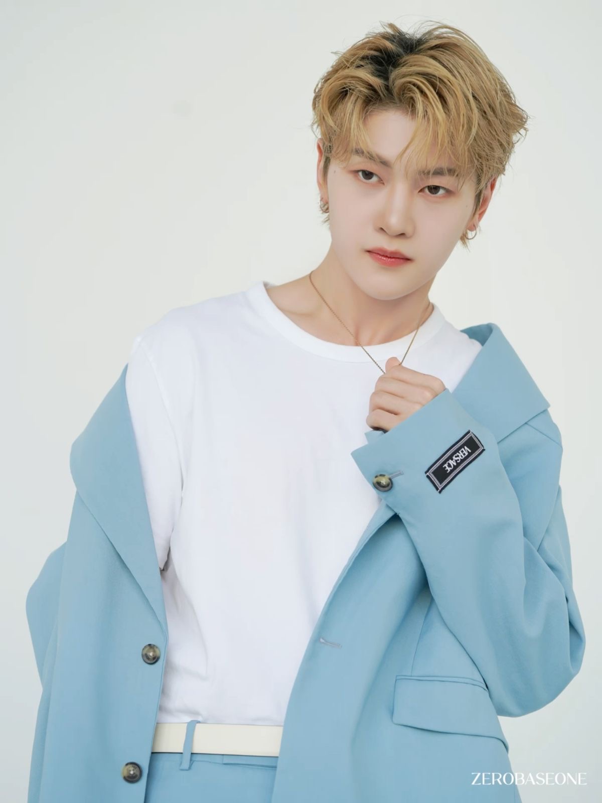 a  portrait of sung hanbin from the kpop group zerobaseone (zb1). he is wearing a pastel blue suit jacket draped over his shoulders and a simple white tshirt. he is accessorized with a silver chain necklace and a white belt.  the background is plain white.