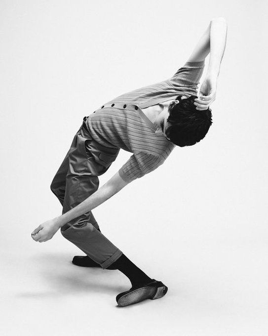 a black and white, fullbody shot of a male model in a contemporary dance pose. he is wearing a ribbed knit shirt and trousers, and is positioned in a dynamic, expressive pose that showcases his flexibility and athleticism. the background is plain and minimalist, allowing the model and his movement to be the focal point. the image is artistic, stylish, and suggests themes of elegance and grace.