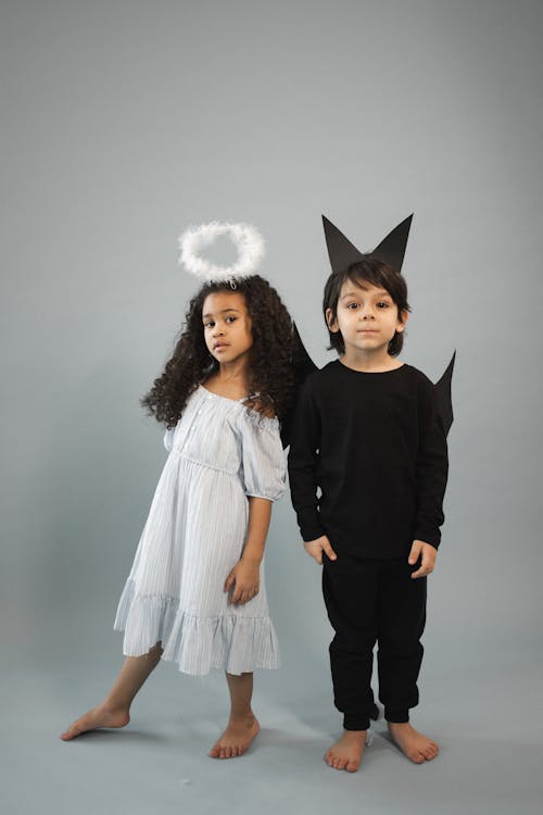 a fullbody shot of a girl and a boy dressed in angel and devil costumes, respectively. the girl is wearing a light blue dress and a halo, while the boy is wearing a black outfit and devil horns. they are standing barefoot on a grey background.  they appear to be siblings or friends, posing playfully for the camera. the image suggests a halloween or themed photoshoot.