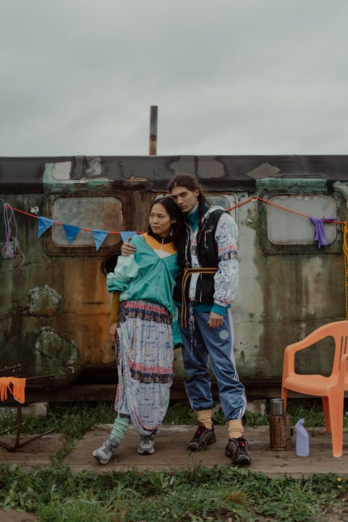 a young couple stands closely together against a backdrop of an old, rusty train car. the woman sports a vibrant turquoise jacket and a patterned skirt, complemented by mint green socks and sneakers. her companion, a man, is dressed in a black and white patterned jacket, blue sweatpants, and black sneakers.  they are both casually dressed, and the overall aesthetic is grunge and slightly vintage. orange plastic chairs and colored bunting add a pop of color to the otherwise muted scene. the background provides a contrast to the couple's stylish clothing. the overall tone is relaxed and stylish.