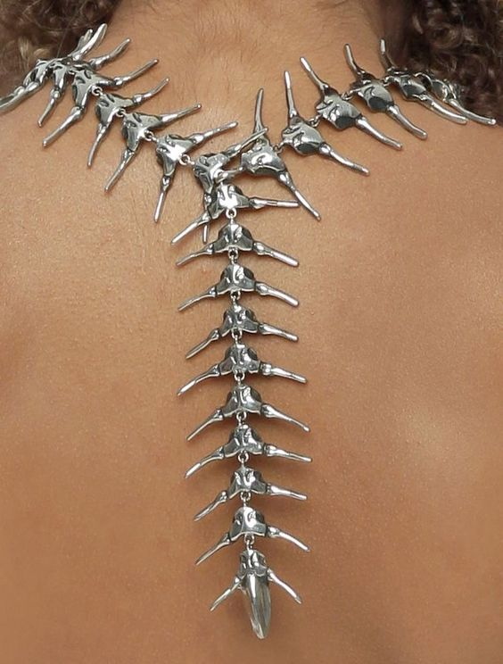 the photo shows the back of a person with brown skin wearing a long silver necklace. the necklace hangs down their back, resembling a spine. each segment is intricately designed and cast in shiny silver. the photo is a closeup, taken in a studio with good lighting.