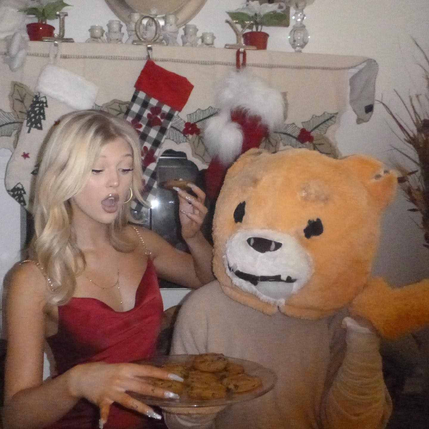 a woman with blonde hair wearing a red dress is holding a plate of cookies and a cookie to her mouth. next to her stands a person wearing a teddy bear costume. the background shows a fireplace mantle decorated with christmas stockings and other decorations.