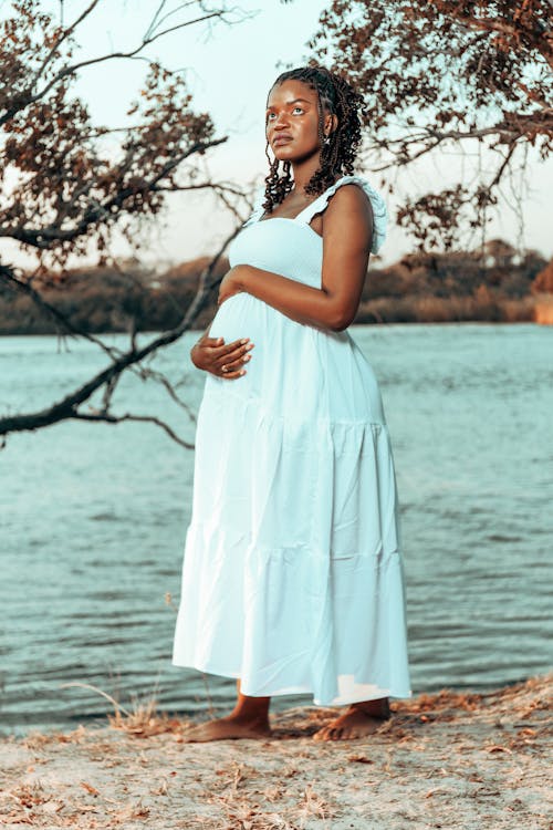 a pregnant black woman in a flowing white maxi dress stands barefoot on the shore of a calm lake.  she's gently holding her belly, and her expression is serene and peaceful.  the setting is idyllic, with a tree in the background and soft sunlight illuminating the scene. the overall mood is one of tranquility and anticipation.