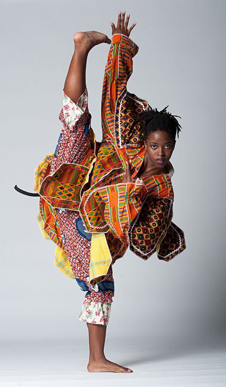 a young woman is standing on one leg, the other leg is stretched straight up in the air. she is wearing a colorful outfit with patterns and fabric that are traditional and beautiful. the woman is looking at the camera with a serious expression. her arm is stretched out in the air and her hand is open, as if she is in middance. the picture is taken in a studio with a white background.
