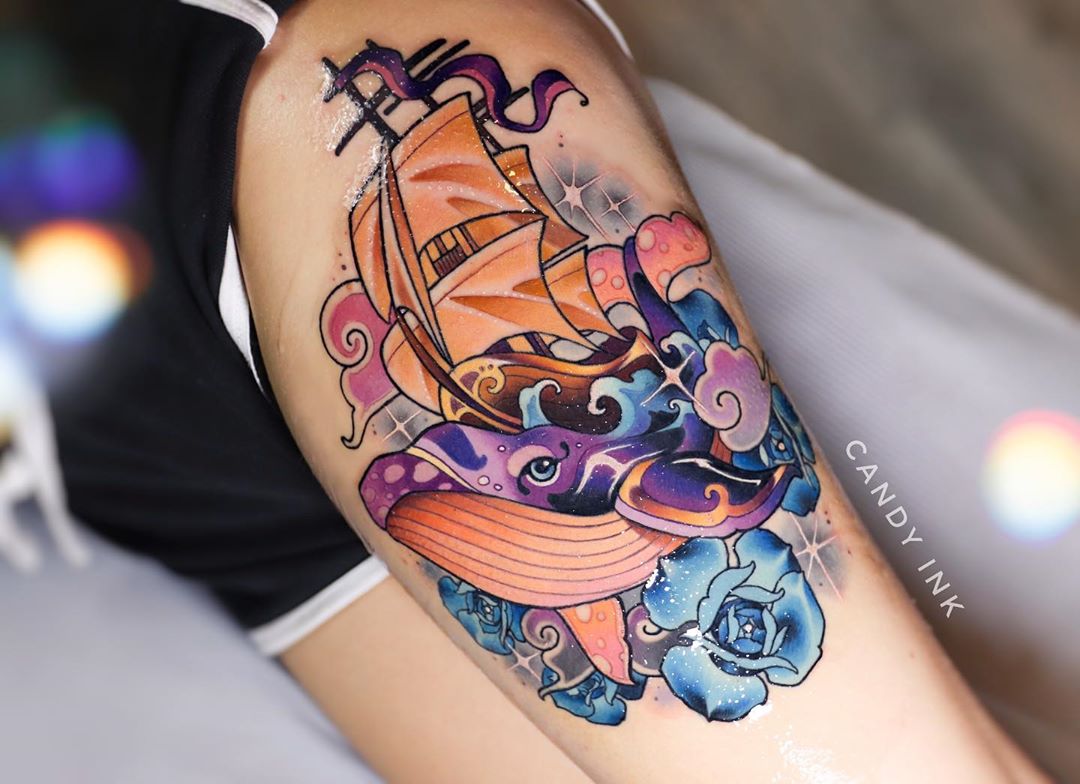 this closeup image showcases a vibrant neotraditional tattoo on someone's upper arm. the tattoo features a large sailing ship perched atop a stylized whale. the whale, rendered in shades of orange, purple, and blue, has a whimsical, almost cartoonish appearance. the ship, with its billowing sails, is depicted in golden yellows and oranges. surrounding the main subjects are stylized waves and blue roses, further enhancing the nautical theme.  the overall effect is whimsical and eyecatching, a testament to the skill of the tattoo artist. the "candy ink" watermark suggests the tattoo parlor or artist's branding.