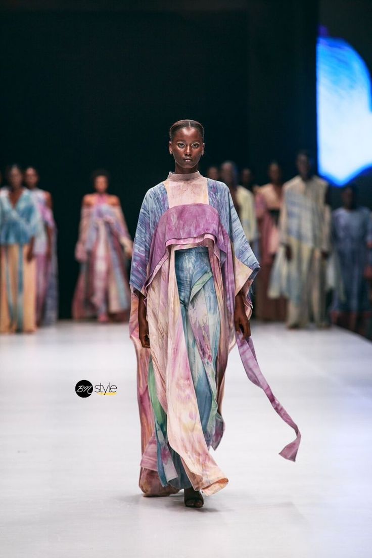 a darkskinned female model walks the runway wearing a long, flowing tiedye dress in pastel colors. the dress has a unique design and features a capelike overlay.  other models wearing similar clothing are visible in the background, blurred, indicating a fashion show. the setting is a runway with a dark background and a partial view of a light blue backdrop. a watermark for "bm style" is present. the overall impression is one of elegance and modern african fashion.