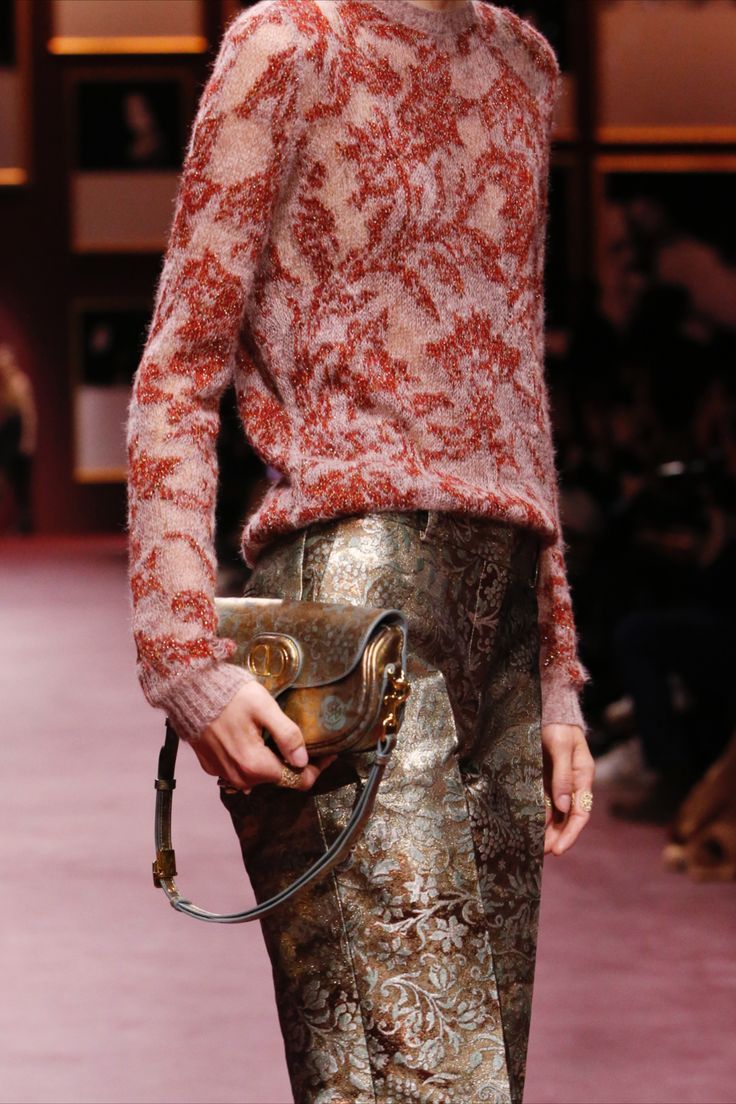 loseup view of a fashion model showcasing a stylish autumn/winter outfit. she's wearing a loosefitting, reddishpink floral patterned knit sweater and metallic gold pants with an intricate floral brocade design. the model carries a small, structured handbag with a metallic finish. the background is blurred, suggesting a runway setting.  the image focuses on the textures and patterns of the clothes and accessories.