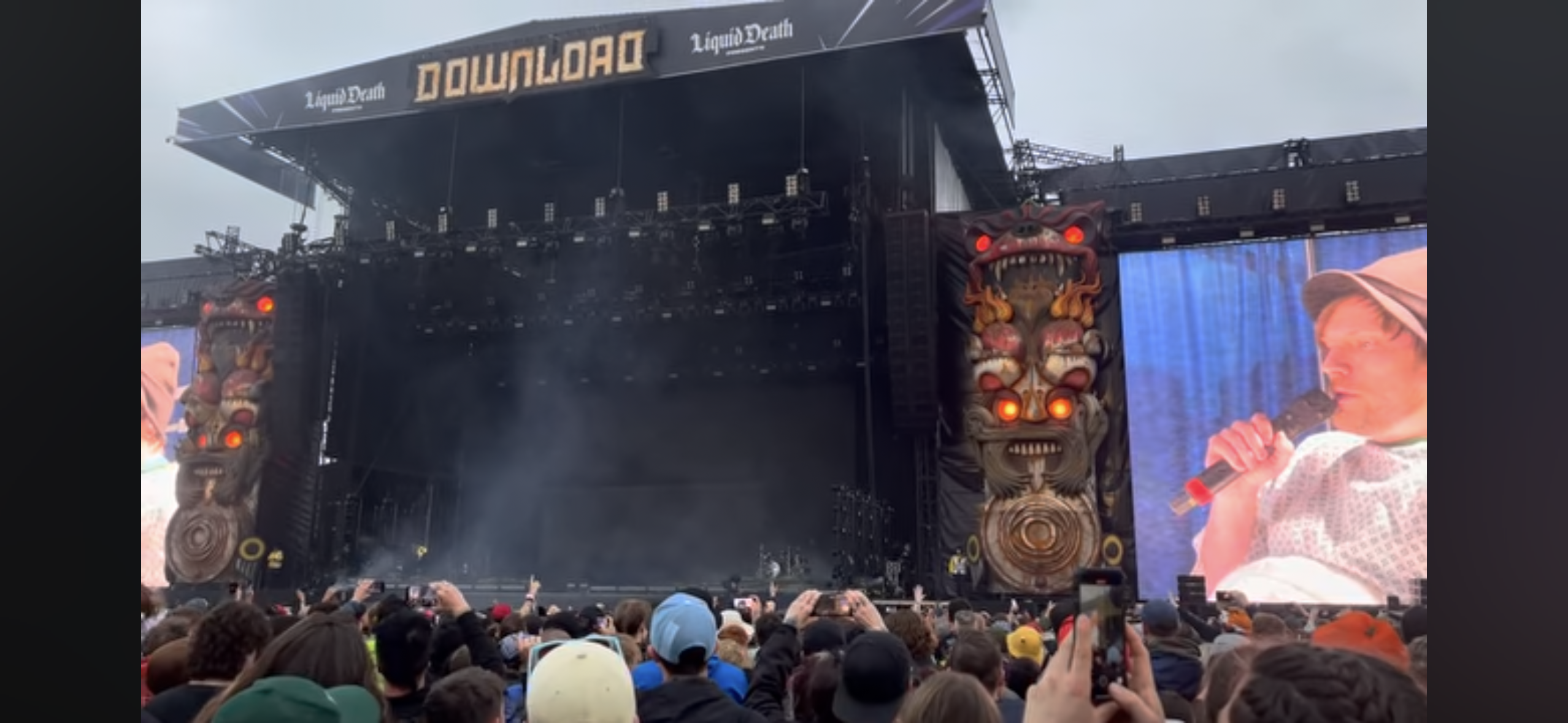 a large crowd of people are gathered at a music festival, facing a stage. the festival is called download and the stage is covered with a giant black sheet. the stage is adorned with large demonic masks. there is a large screen on the right side of the stage showing a singer holding a microphone. the crowd is excited and the atmosphere is electric.