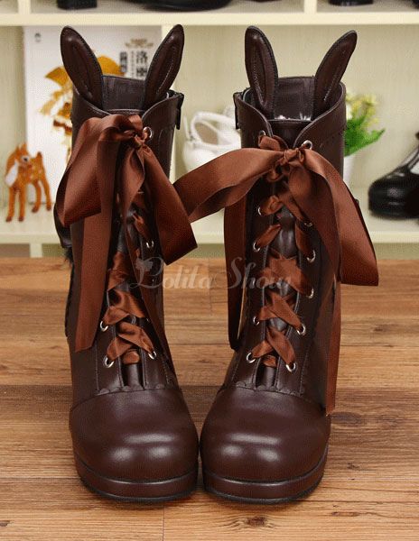 a closeup shot of two brown boots on a wooden surface. the boots have bunny ears attached to the top and are adorned with large brown ribbons. the boots have a platform sole and are laced up with brown laces. the style of the boots is associated with lolita fashion. there are shelves with other shoes and decorative objects in the background.