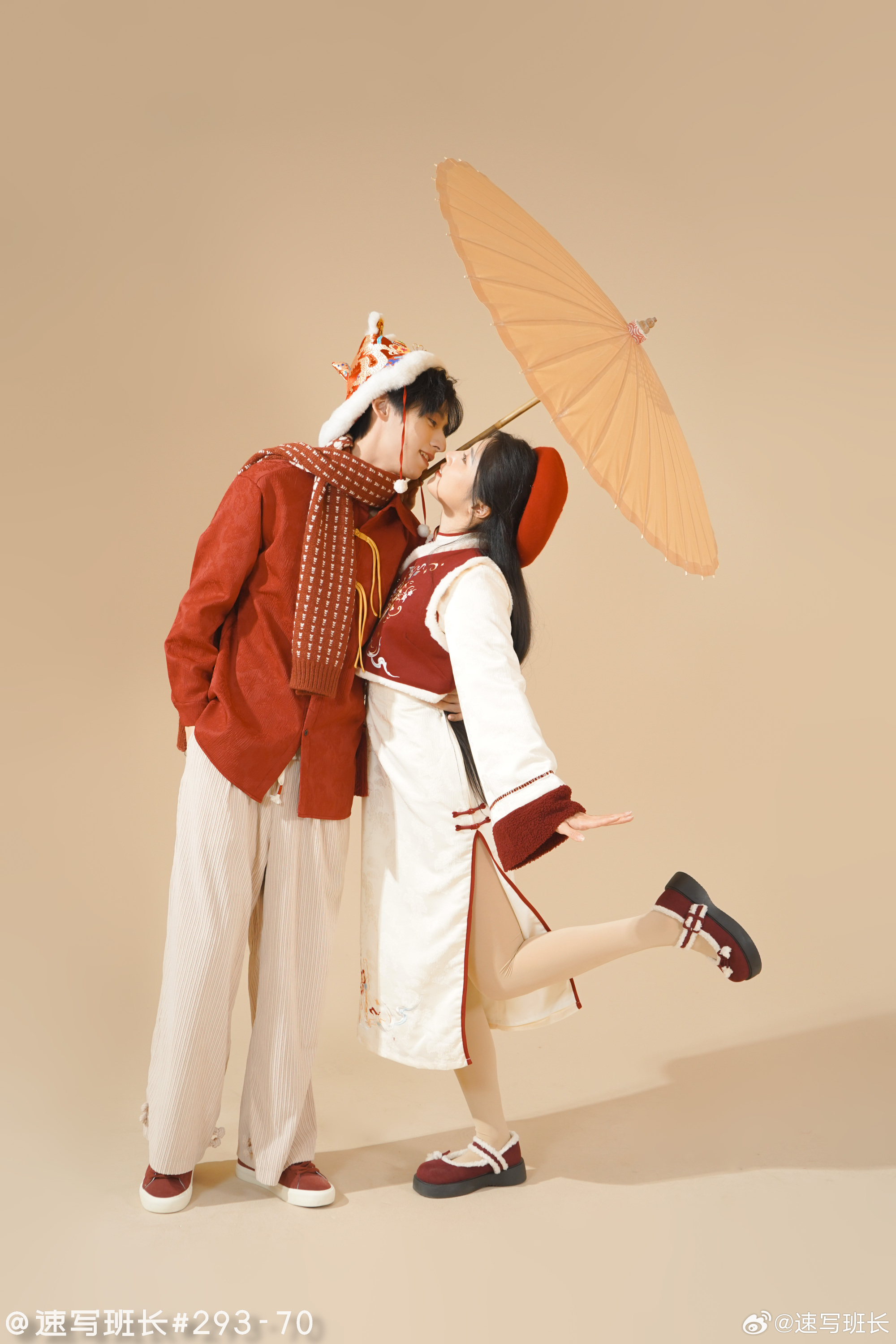 a man and a woman are posing for a picture in what appears to be a studio. they are wearing traditional chinese new year clothing in red, white, and gold. the man is wearing a red jacket with gold detailing, a white scarf, white pants with a pattern of red stripes, a gold crown, and white shoes. the woman is wearing a white dress with red details, a red hat, and black shoes. the man is holding a paper parasol over the couple, and they are both smiling and looking at each other. the background is a light brown color.  the image has chinese characters at the bottom left and right.