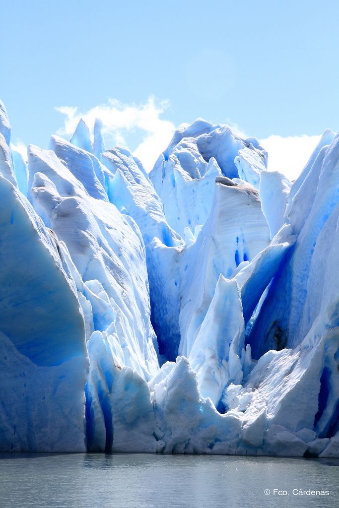 a closeup shot of a glacier with striking blue hues meeting the calm water below. the glacier is textured with various formations and crevasses, set against a bright sky with faint clouds.