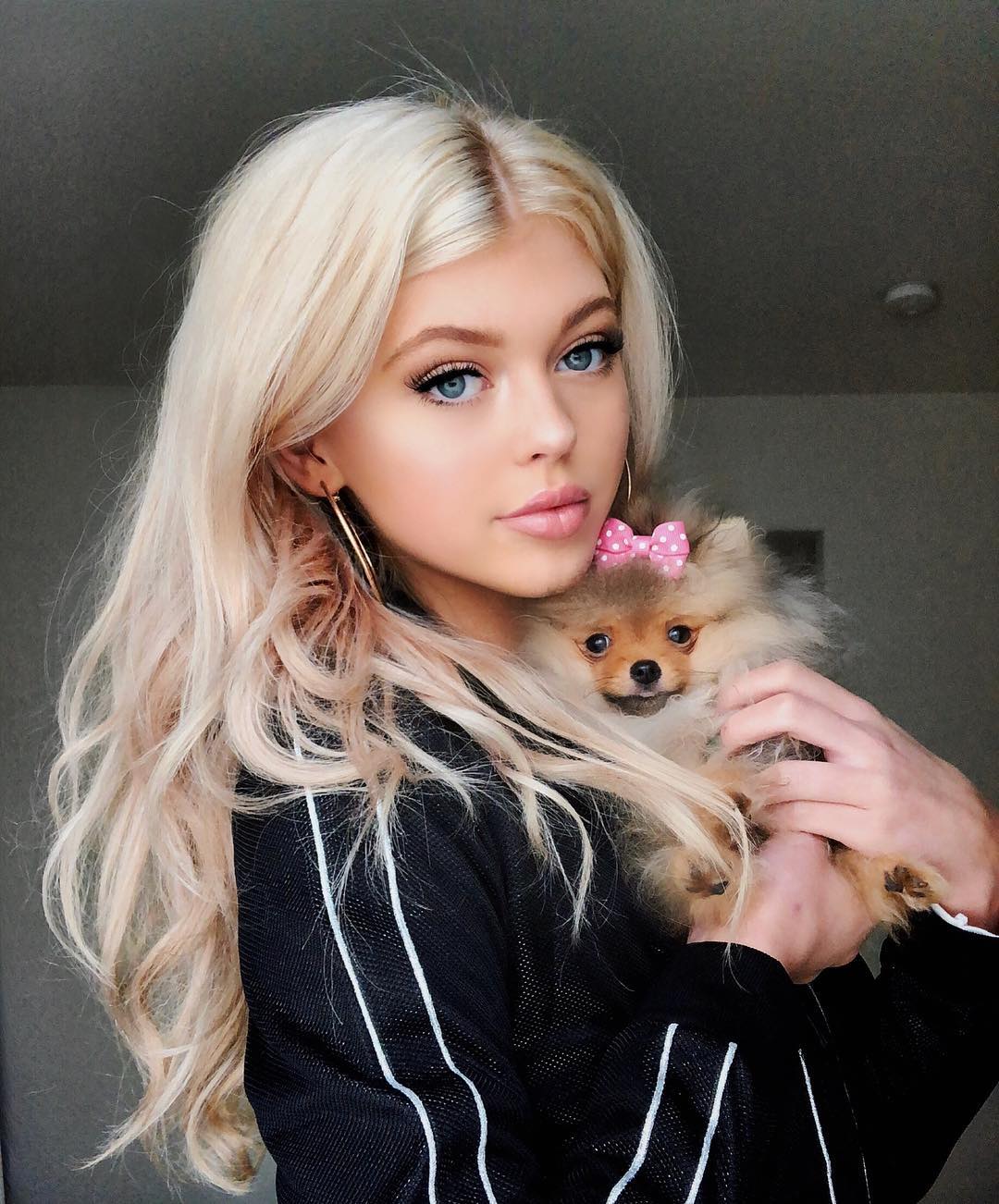a young woman with long blonde hair and blue eyes is holding a fluffy pomeranian dog. the dog has a pink bow on its head. the woman is wearing a black jacket with white stripes and hoop earrings.
