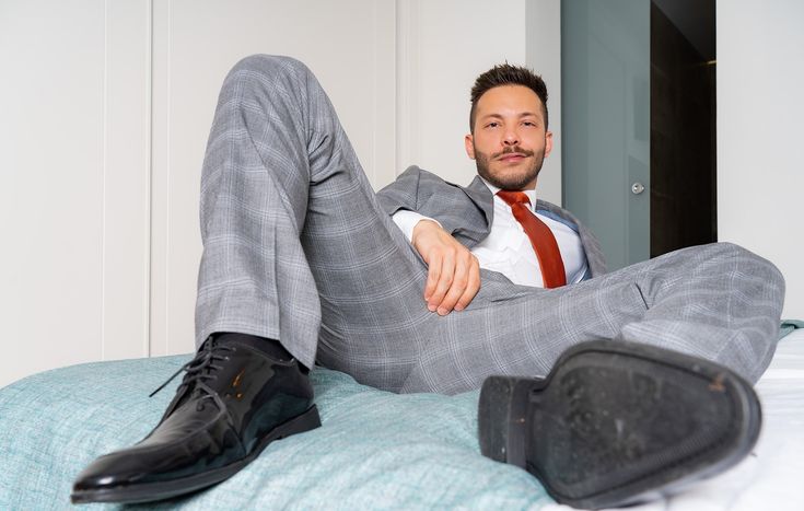 a man in a grey suit and red tie sits on a bed with his legs crossed. his black shoes are visible in the foreground. he is looking confidently at the camera. the image conveys an air of confidence and sophistication.