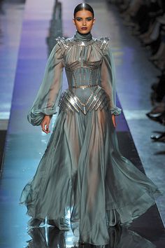 a female model walks the runway in a long, flowing gown. the gown is made of a light blue and silver fabric, and it is adorned with sequins and other embellishments. the model is wearing her hair up, and she has a glamorous makeup look. the background is blurred, and there are other people walking around in the background.