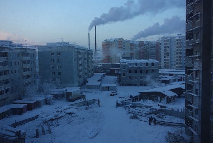 a snowy city scene with tall buildings and a smokestack in the distance. the photo is taken from a nearby apartment building, looking out over the city. there is a light dusting of snow on the ground and the buildings are mostly grey and white. the sky is a pale blue, with some clouds and fog. there is a car driving down the street in the distance and it looks like it is snowing lightly. the buildings in the background look like they are in various stages of construction. there is scaffolding on the building to the right of the frame. there are also some trees covered in snow.