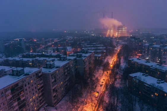 an aerial, highangle, long shot of a city at night during a winter snowfall. the image is taken at twilight, with a dusky purplishblue sky. the city is composed mostly of apartment buildings covered in snow, with streetlights illuminating the roadways and creating warm orange hues along a treelined street. in the distance, a large power plant with cooling towers is visible, emitting smoke or steam, adding to the atmospheric quality of the image. the overall feel is one of coldness and quiet intensity, with the lights of the city contrasting against the dark sky and snowy landscape.
