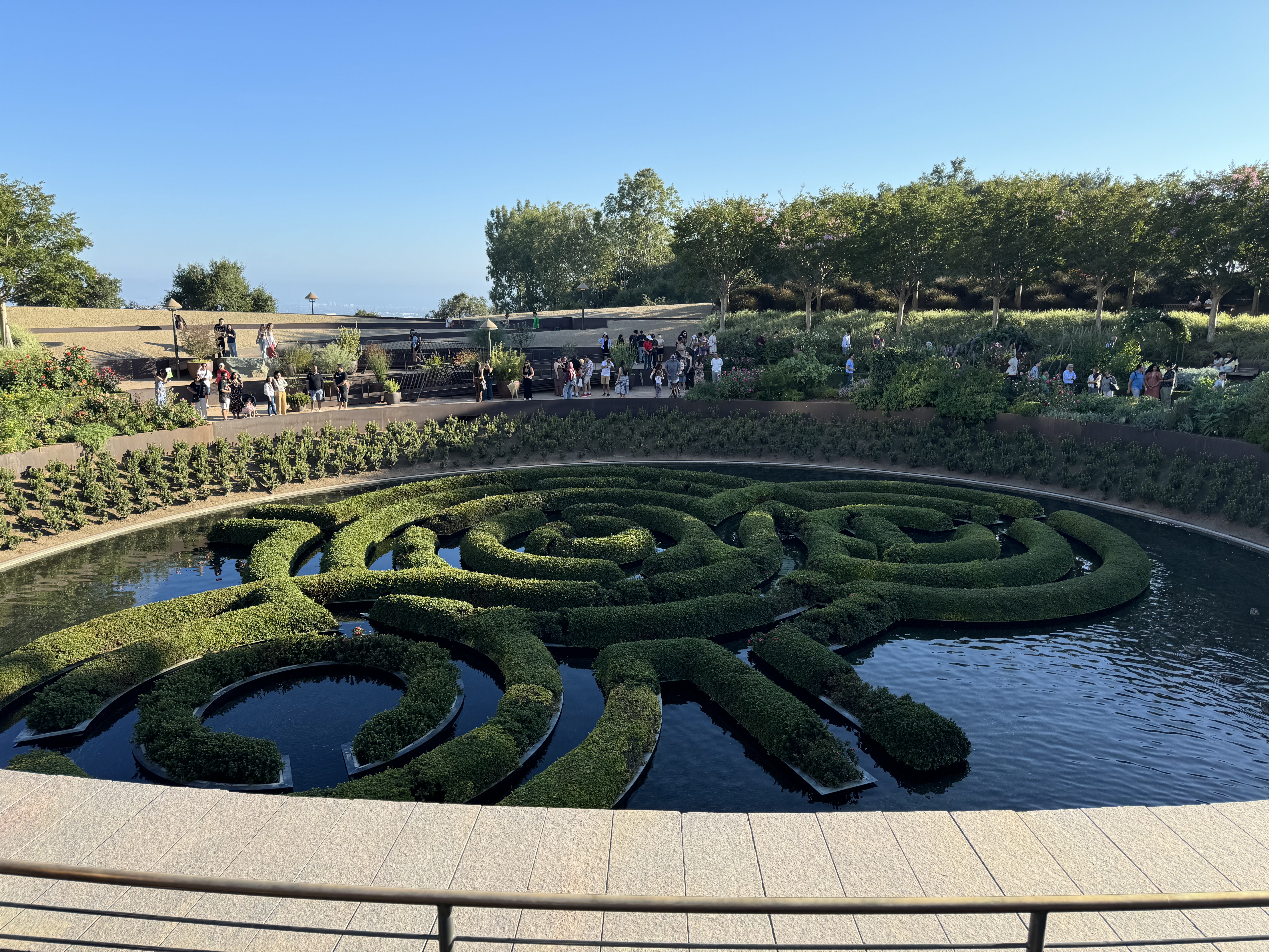 a highangle view of a circular fountain designed as a maze. the maze is made of precisely trimmed green hedges, with pathways of water separating the sections. the fountain is surrounded by a paved walkway and a variety of trees and shrubs. in the background, there are more trees and a clear blue sky. the scene is bustling with people who are admiring the fountain and enjoying the sunny weather.