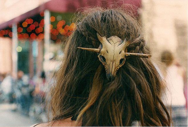 a closeup image of a woman's back, her long brown hair is styled and secured with a deer skull hair clip.  the image is taken outdoors in a city setting, with people in the background blurred.