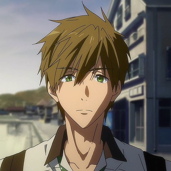 the image shows a young man with brown hair and green eyes. he is standing outdoors on a street with buildings in the background. the lighting is daylight and the perspective is eye level.