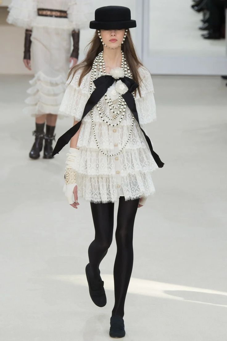 a woman is walking down a runway. she is wearing a white lace dress with black ribbon accents and pearl necklaces. she is wearing black gloves, a black hat, and black shoes. she is also wearing black tights. the runway is white and there is a white backdrop behind her. she is posed as if she is walking down a runway.