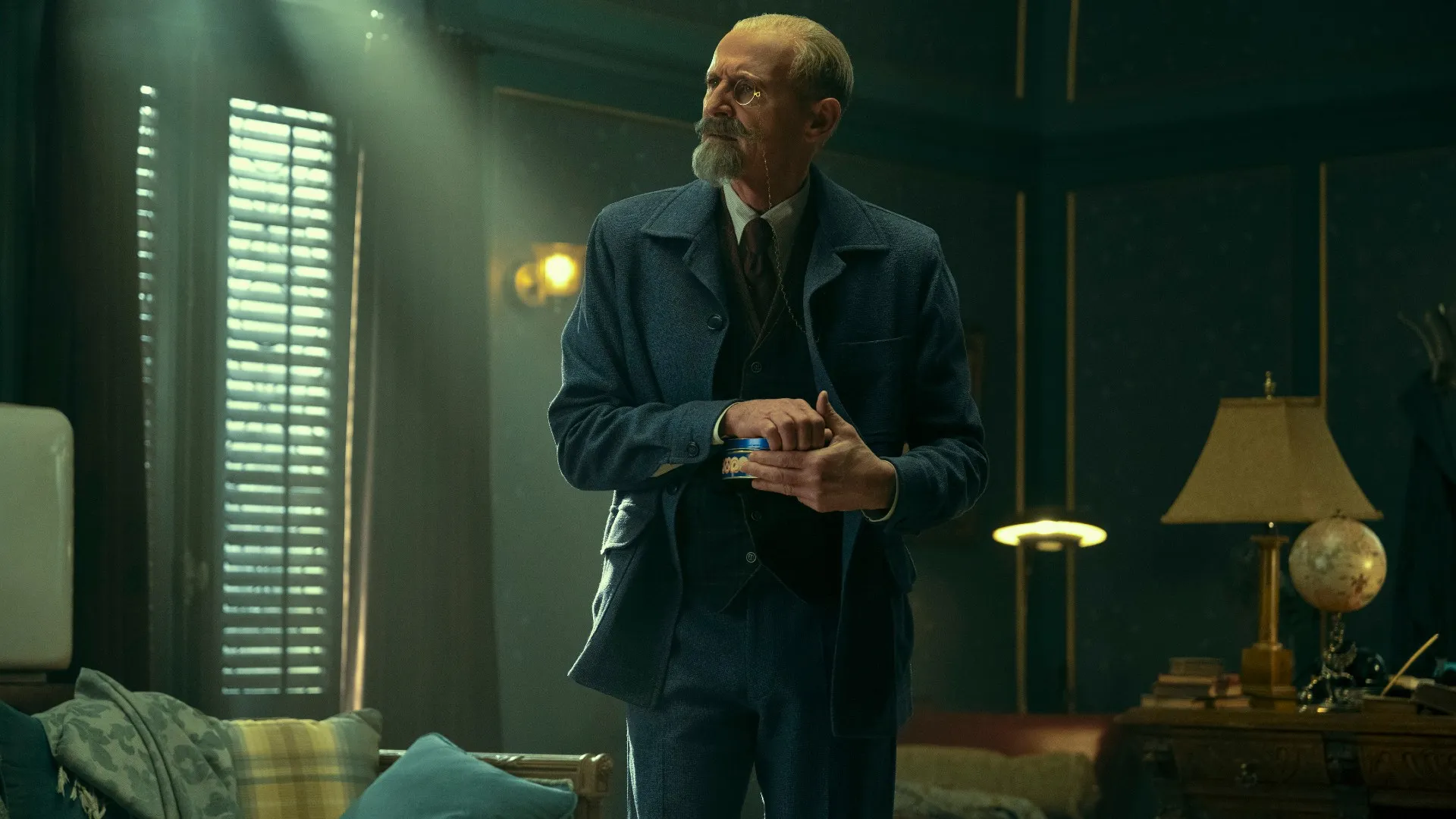 an older man with a distinguished grey beard and dressed in a suit is standing in a dimly lit room. he is holding a small, unidentified object in his hands and appears lost in thought. the room has a vintage feel with dark wood paneling and a globe on a side table.  a window with blinds is to the man's left and a lamp is on a table to his right.
