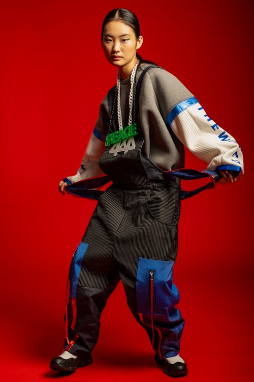 a young asian woman is modeling a stylish outfit against a vibrant red background. she's wearing a grey sweatshirt with blue and white detailing on the sleeves, black and blue overalls with cargo pockets, and chunky black shoes.  she also has on a necklace with a "peace 444" pendant. the overall style is streetwearinspired, contemporary, and trendy. the image is a professional studio shot, focusing on the model and her outfit.  the model is posing dynamically, adjusting her overalls.