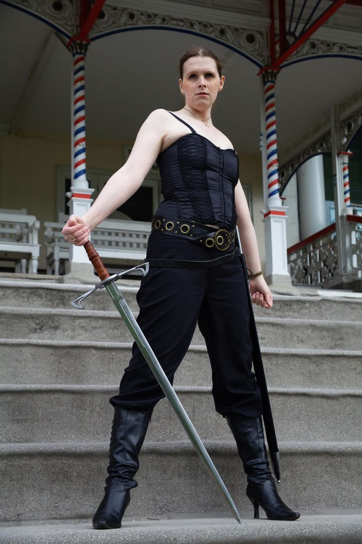 a woman in black clothing stands on a set of stairs holding a sword in front of her. she is posing with her body turned to the right but looking towards the camera. she appears to be in a warrior or fantasy type of pose.