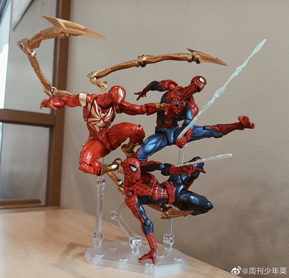 the image shows three spiderman action figures in a dynamic pose. one is the classic red and blue spiderman, another is the scarlet spider, and a third is a red and gold version with mechanical spider arms. the figures are posed on clear stands to simulate them swinging through the air. they are placed on a table with a window in the background.