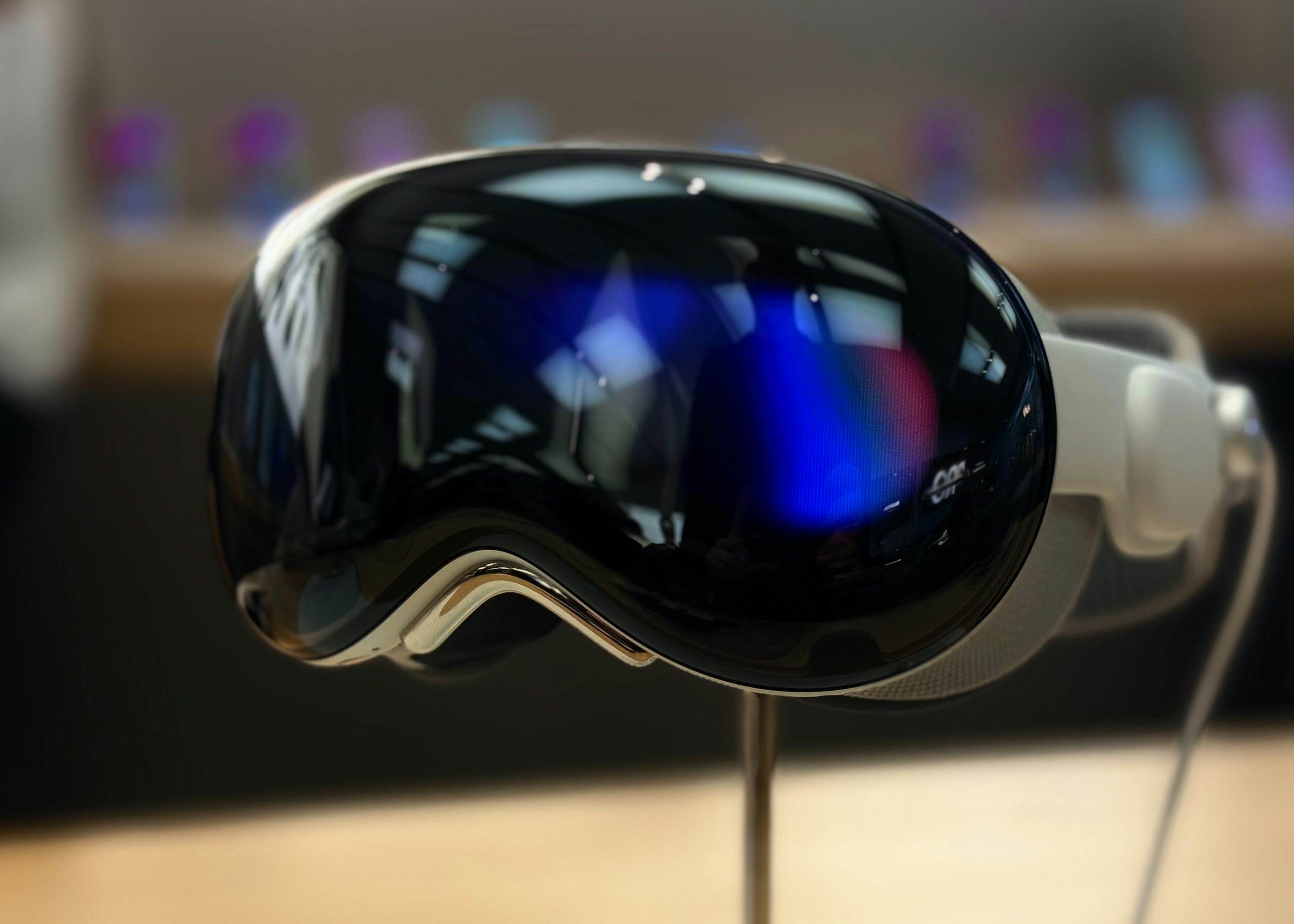 a closeup shot of the apple vision pro vr headset on display. the headset features a sleek black visor with reflections and a white head strap. the device is mounted on a metal stand.